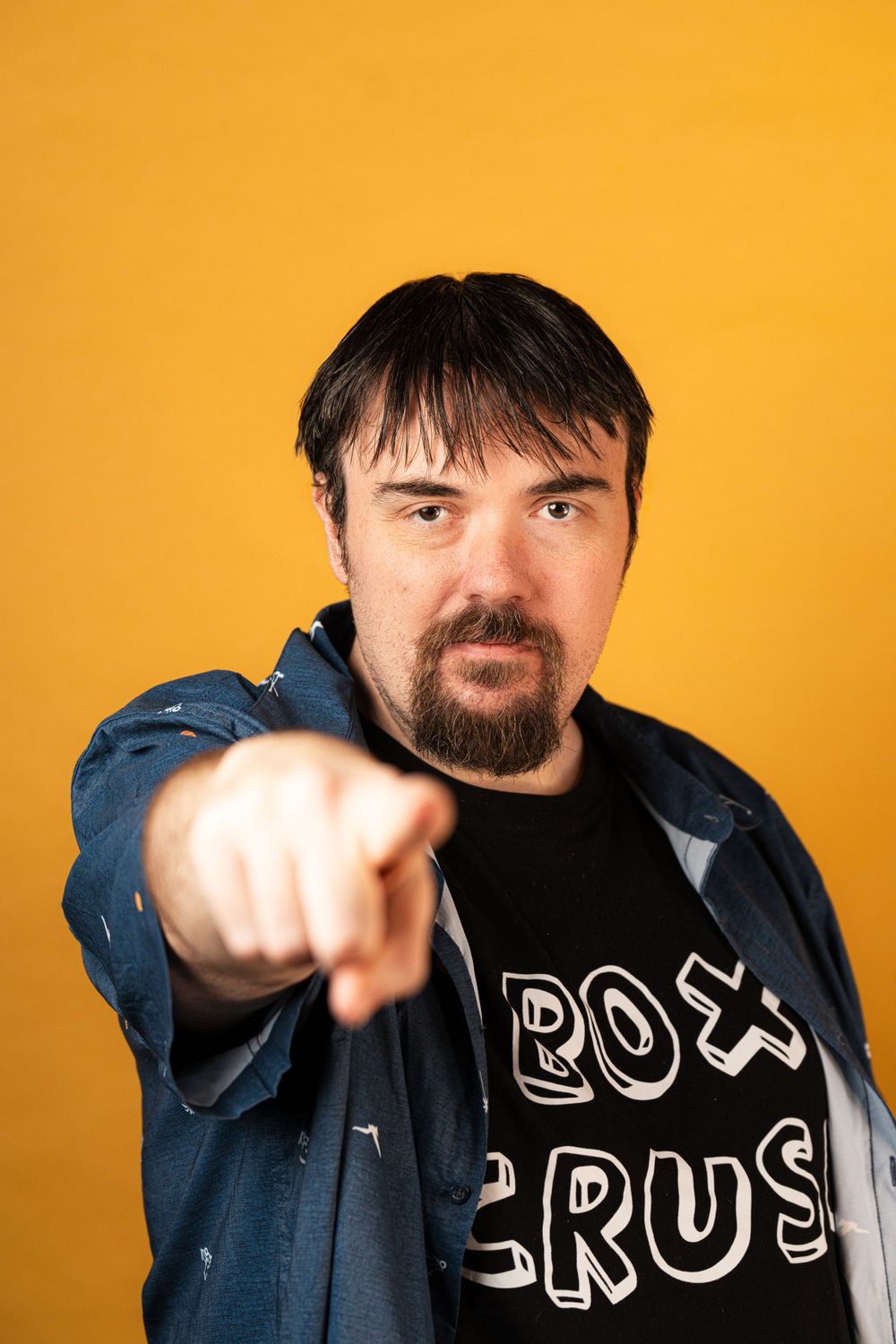 Against an orange background, someone in a denim jacket and band t-shirt points assertively at the camera.