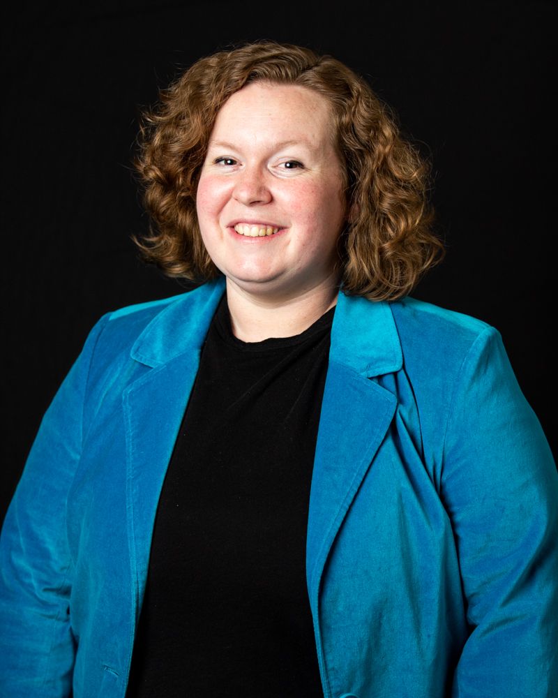 Professional studio portrait showing someone wearing a bright turquoise blazer over a black shirt against dark background.