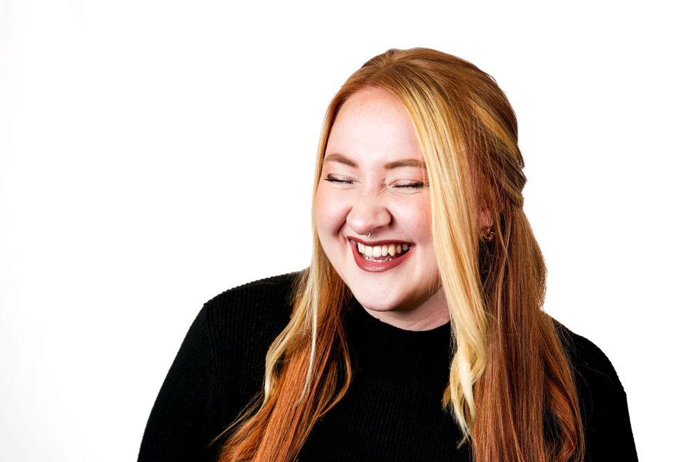 A person with long red hair wearing black laughs joyfully against a white background.