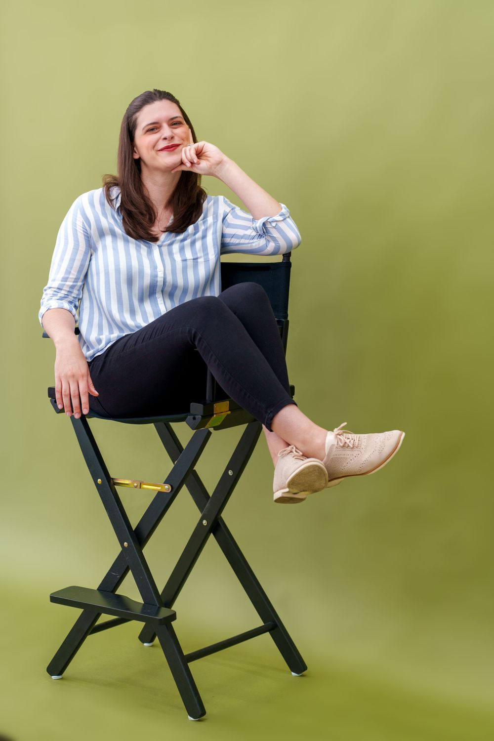 Someone in a blue striped shirt and black pants sits casually on a tall black folding chair against a green backdrop.