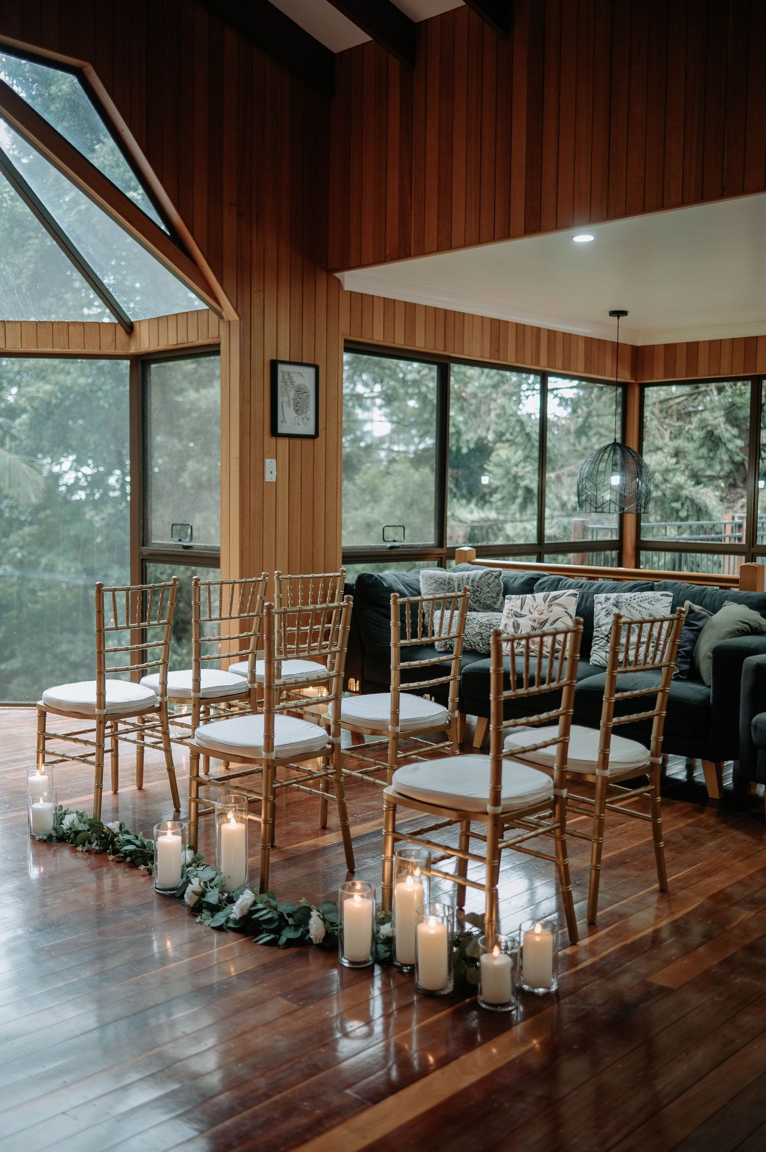 Wedding ceremony setup with wooden chairs and candle-lined aisle in a rustic room with large windows.