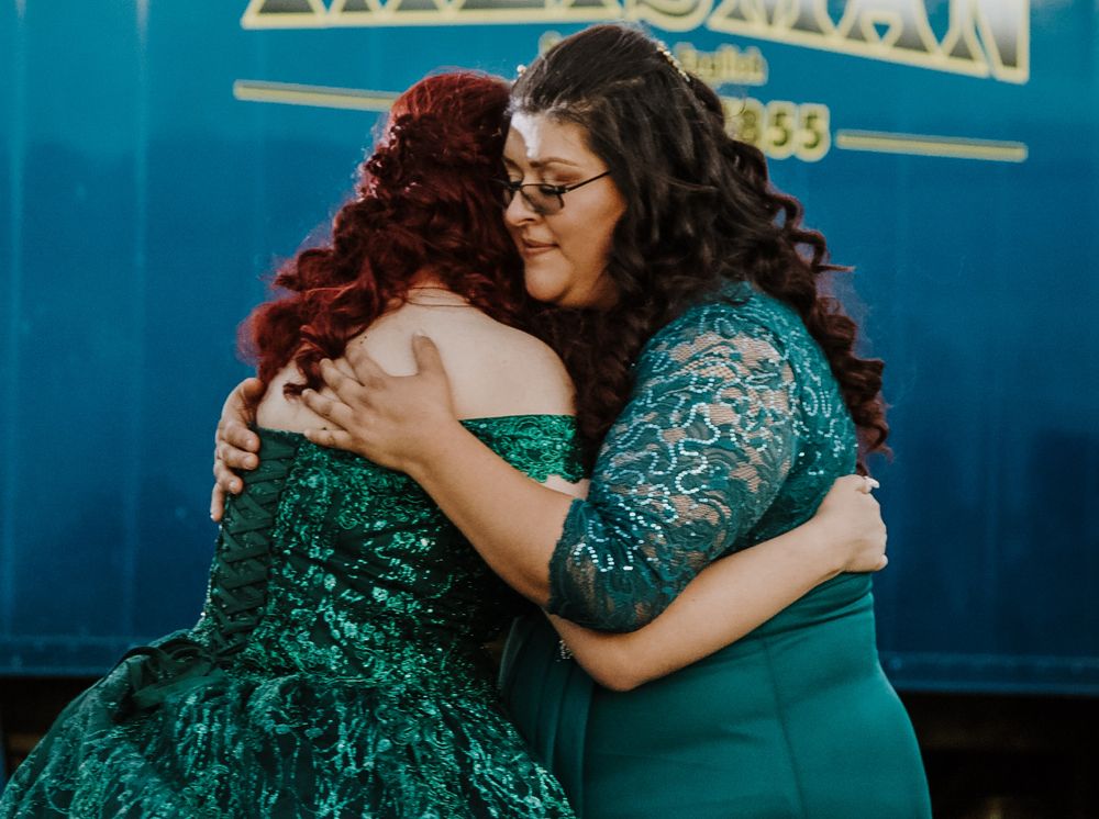 Emotional hug between quinceañera girl in green sequin ballgown and woman in emerald lace dress at outdoor celebration.
