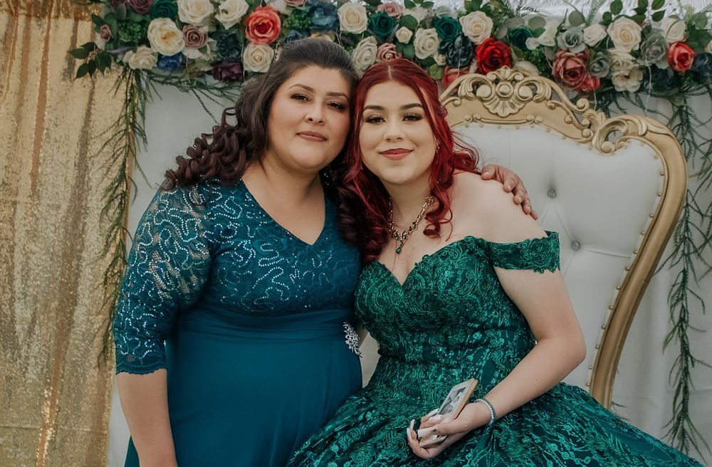 Two women in emerald green dresses pose together in front of a floral backdrop at a quinceañera.