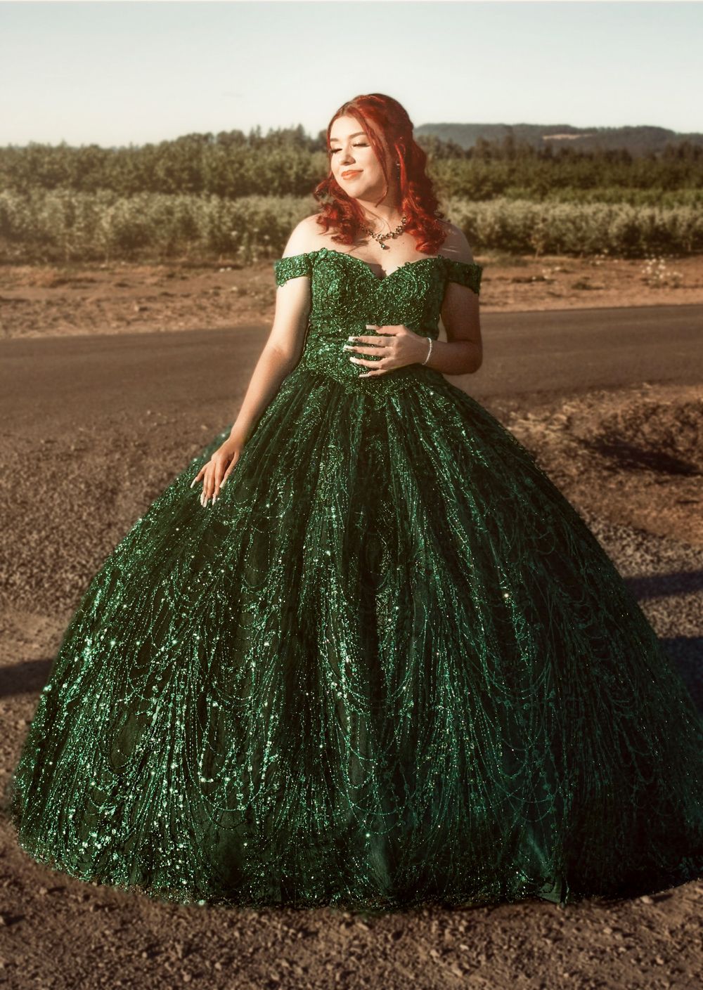 Young woman in breathtaking emerald green sequin off-shoulder quinceañera ballgown posing on rural road at golden hour.