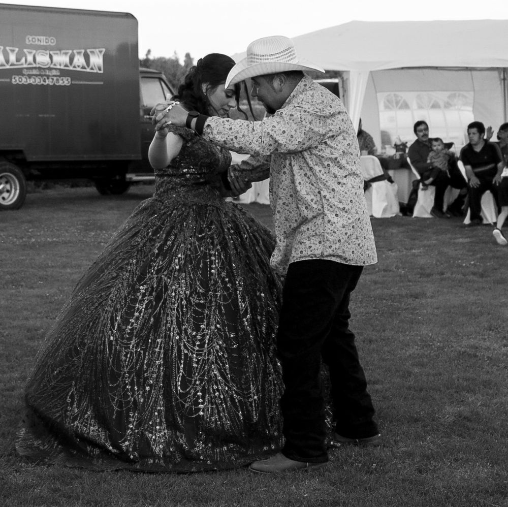 Man in white hat dances with young woman in ornate ball gown at outdoor quinceañera, black and white.