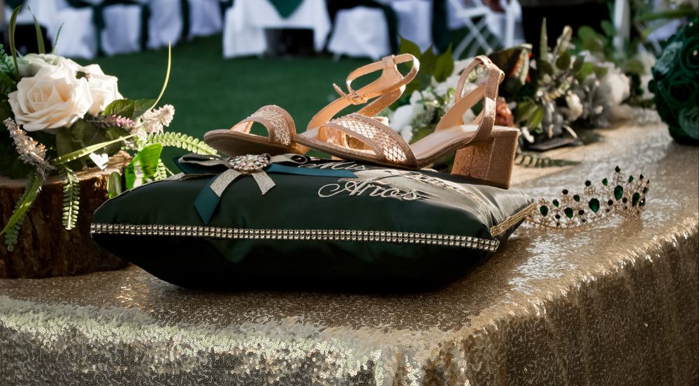 Rose gold heeled sandals and a tiara displayed on a green embroidered quinceañera pillow on a table.