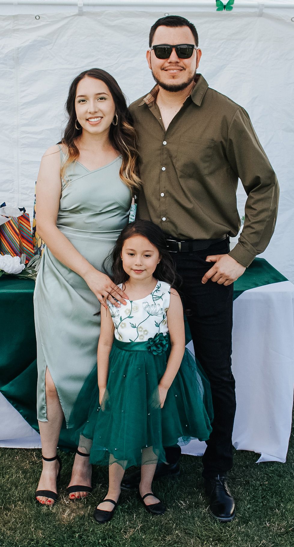 A couple in sage and olive formal wear poses with a young girl in a green tulle dress at an event.