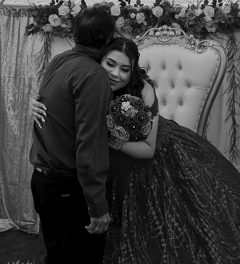 A young woman in a black lace quinceañera gown embraces a man while holding a bouquet at a decorated event.