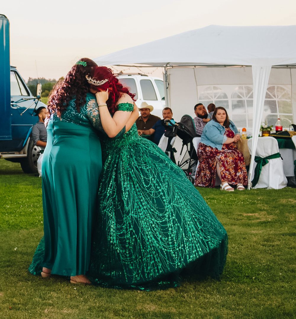 Two women embracing and dancing at quinceañera, both wearing emerald green dresses with guests seated under white tent.