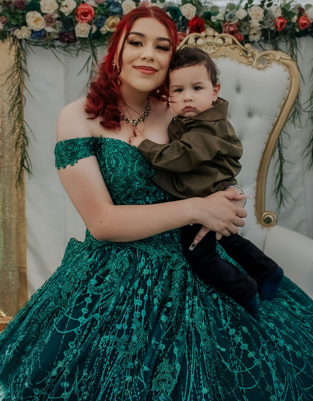 A young woman in a stunning emerald green quinceañera ball gown holds a baby boy on a throne chair.