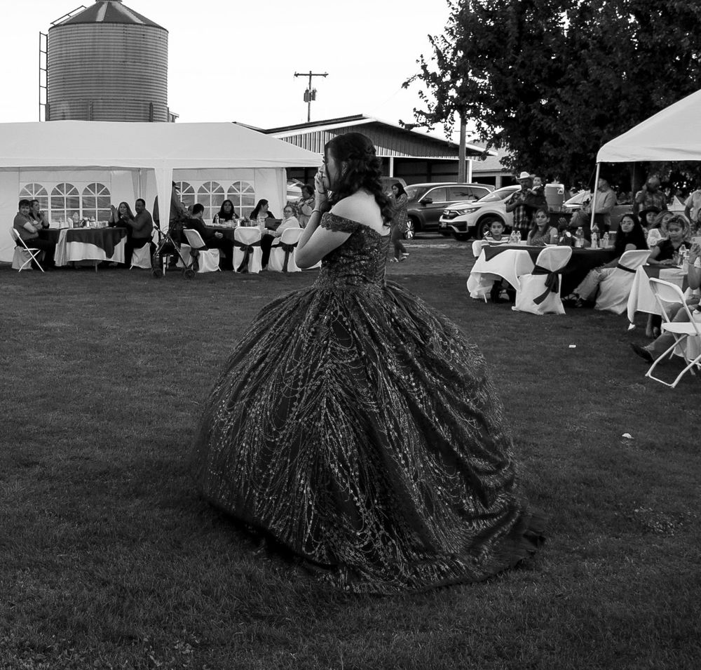 Young woman in elaborate ball gown stands on grass at outdoor quinceañera celebration, black and white.