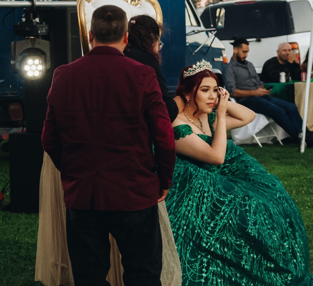 Quinceañera girl in emerald sequin ballgown and tiara stands with escort in burgundy velvet suit at outdoor celebration.