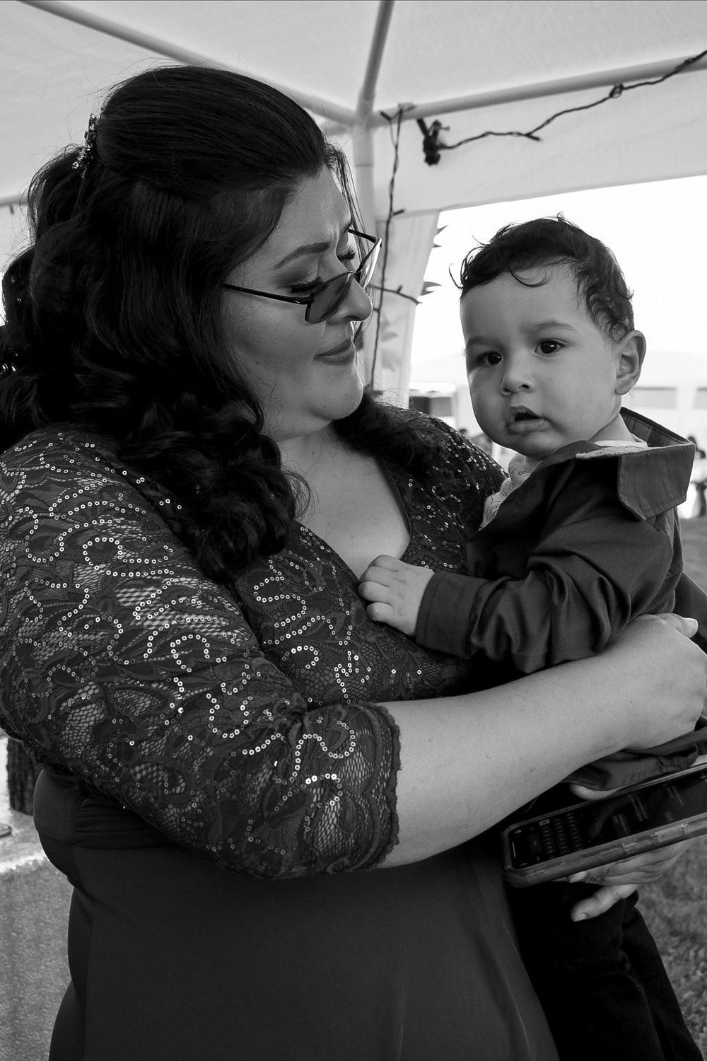 Woman with glasses in lace dress tenderly holds a baby boy in a suit, black and white photo.