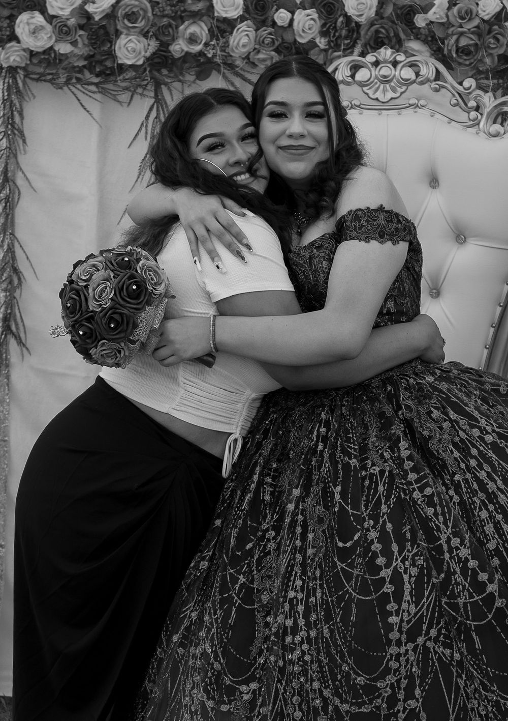 Two young women in formal gowns share a warm hug at a quinceañera celebration, black and white.
