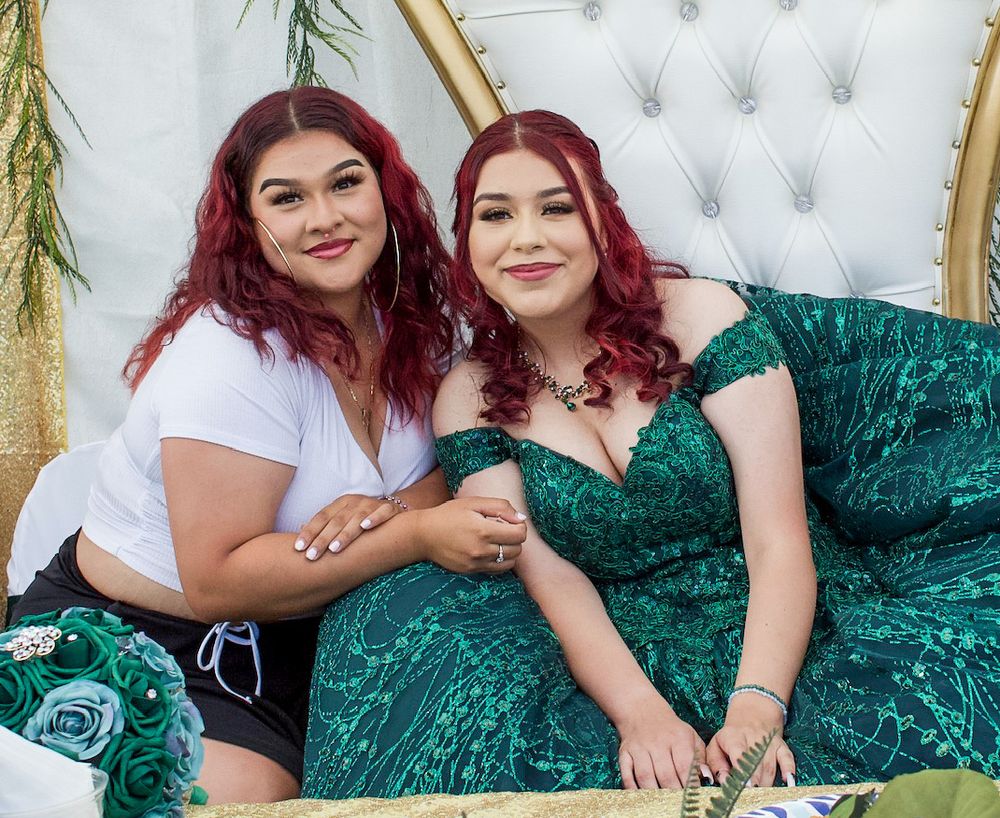 Two women with red hair smile together on a white tufted chair, one wearing a green sequin dress.