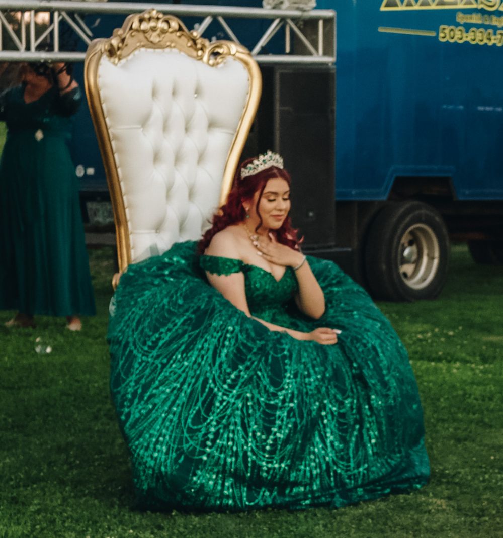Quinceañera girl in stunning emerald green sequin ballgown seated on ornate white and gold throne chair wearing tiara.