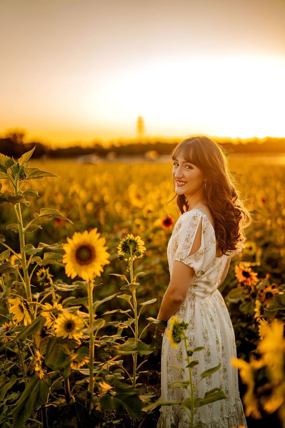 A white dress glows in golden sunlight among blooming sunflowers during a magical summer sunset.
