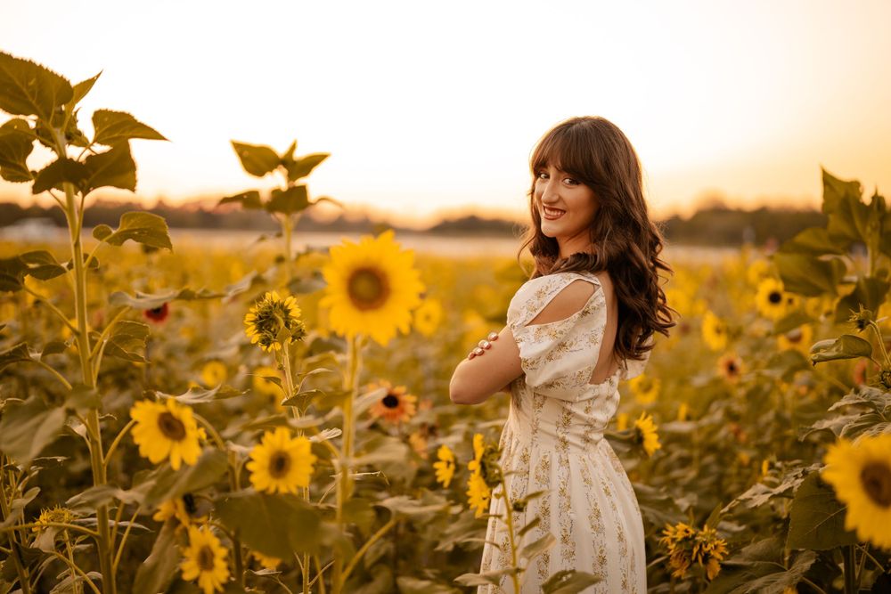 A woman in a white dress stands among blooming sunflowers during golden hour sunset in a summer field.