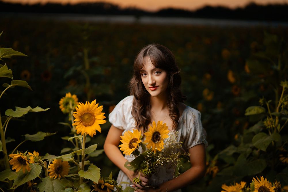 A figure stands in a sunflower field at sunset holding a bouquet of bright yellow sunflowers.