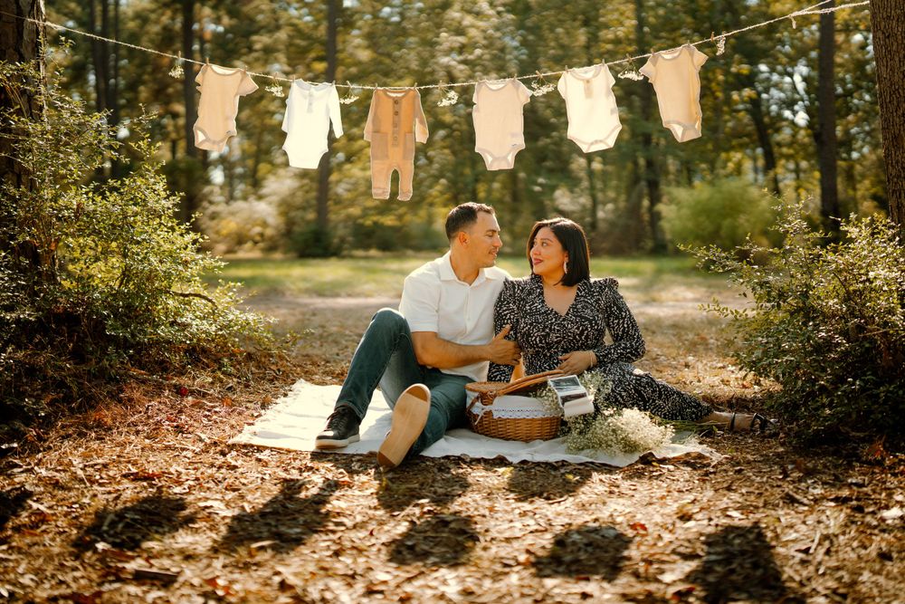A couple sits on a blanket in a forest setting with baby clothes hanging on a clothesline above them.