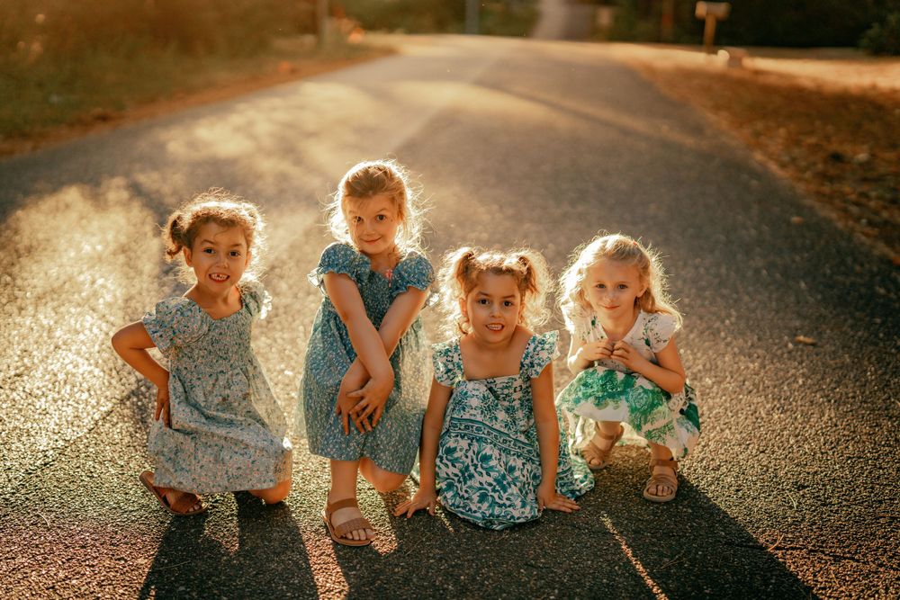 Four young children in summer dresses stand together on a sunlit country road during golden hour.