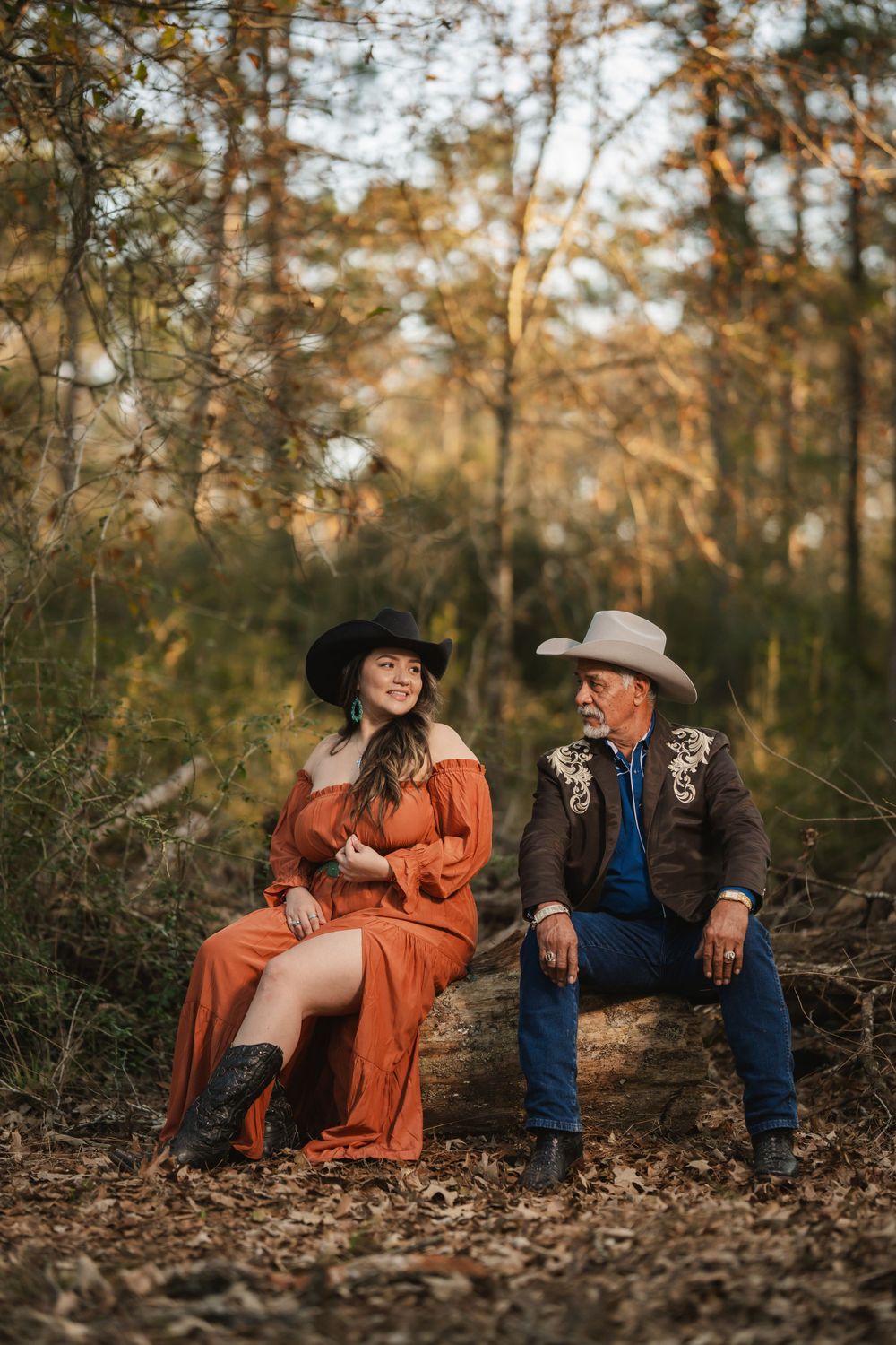 A couple in western attire sits together on logs in a wooded area during golden hour.