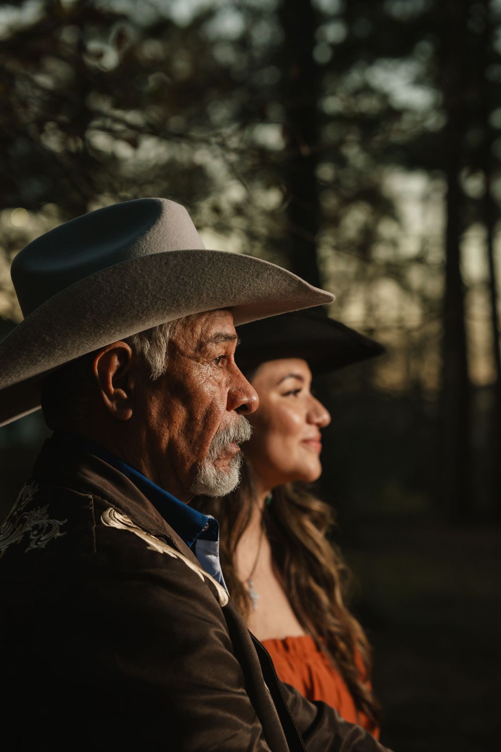 Side profile portrait of two people wearing western cowboy hats in warm sunset lighting through trees.