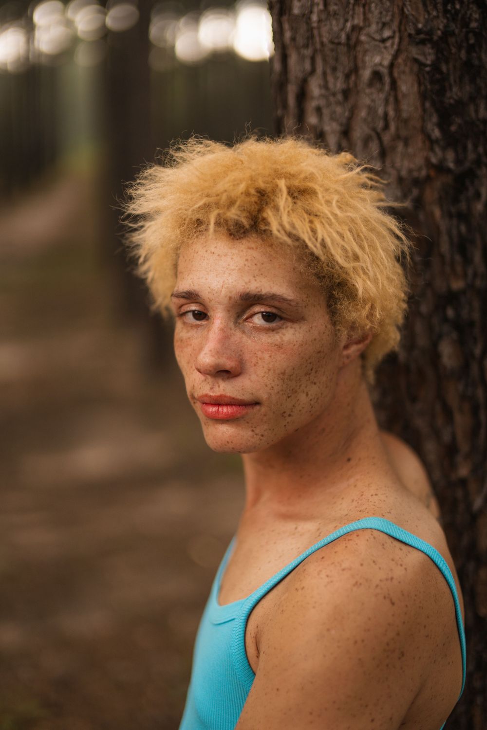 A portrait of someone with bright blonde curly hair wearing a turquoise tank top leaning against a tree trunk.