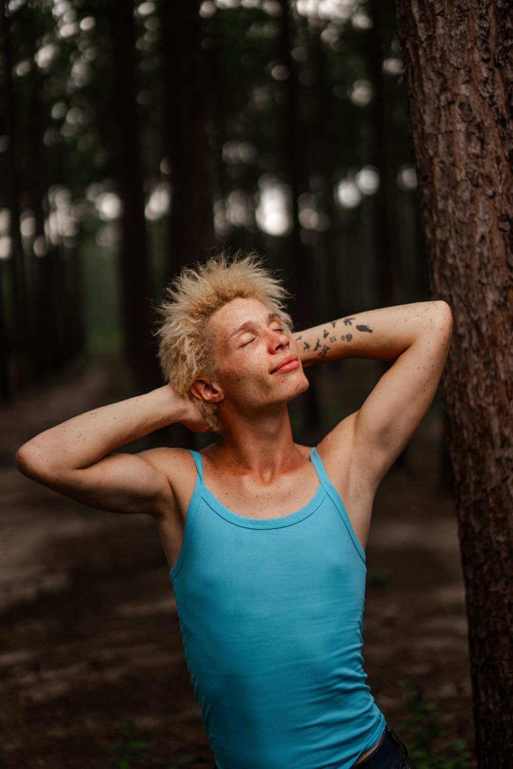 Person in bright turquoise tank top relaxing against a tree trunk in a moody forest setting at dusk.