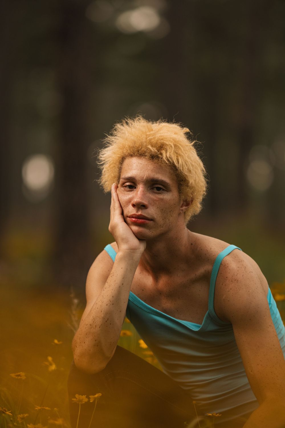 A person with blonde spiky hair wearing a turquoise tank top poses thoughtfully in a moody outdoor setting.