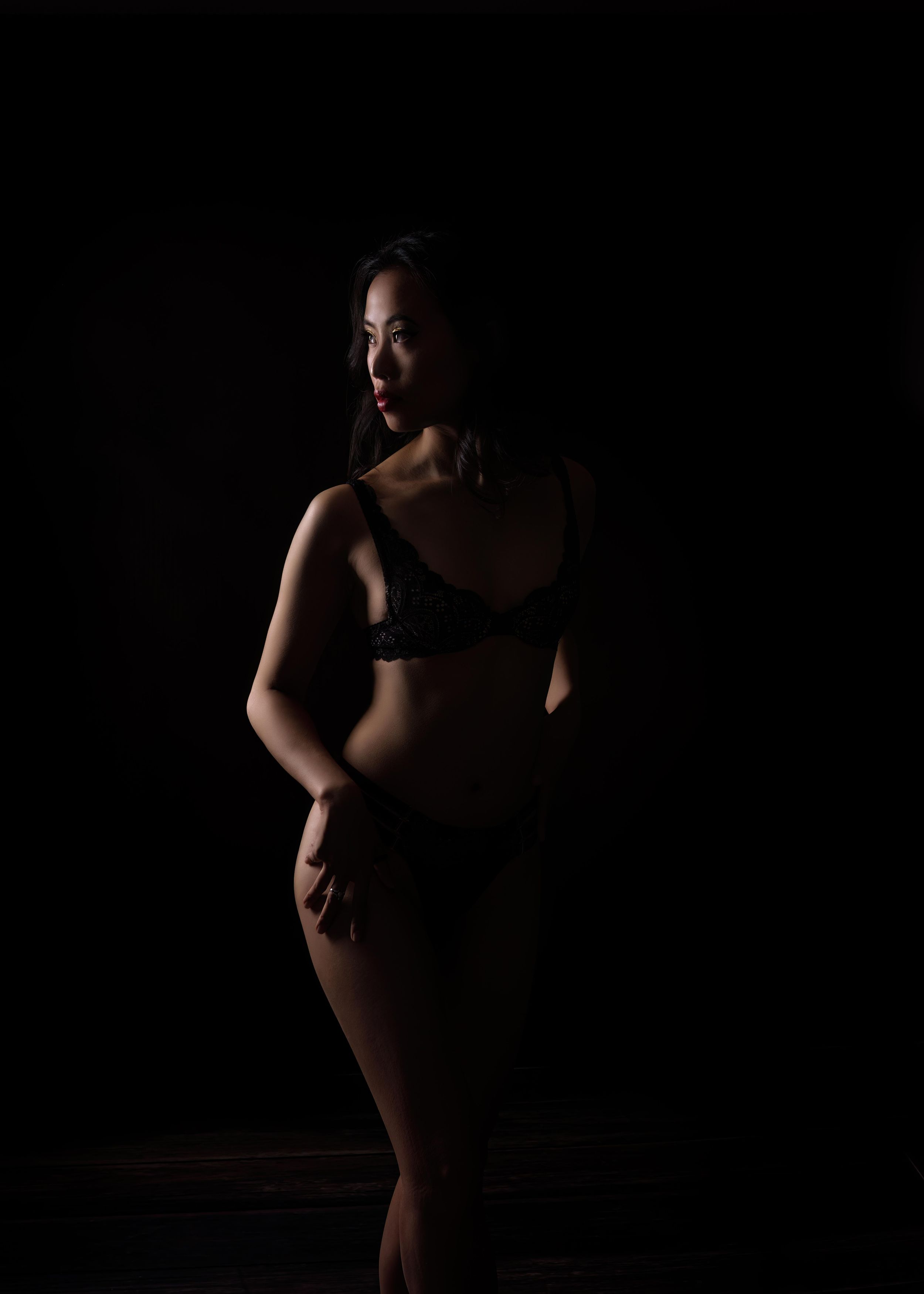 A silhouette of someone in dark lighting stands in an artistic pose wearing black undergarments against a dark background.