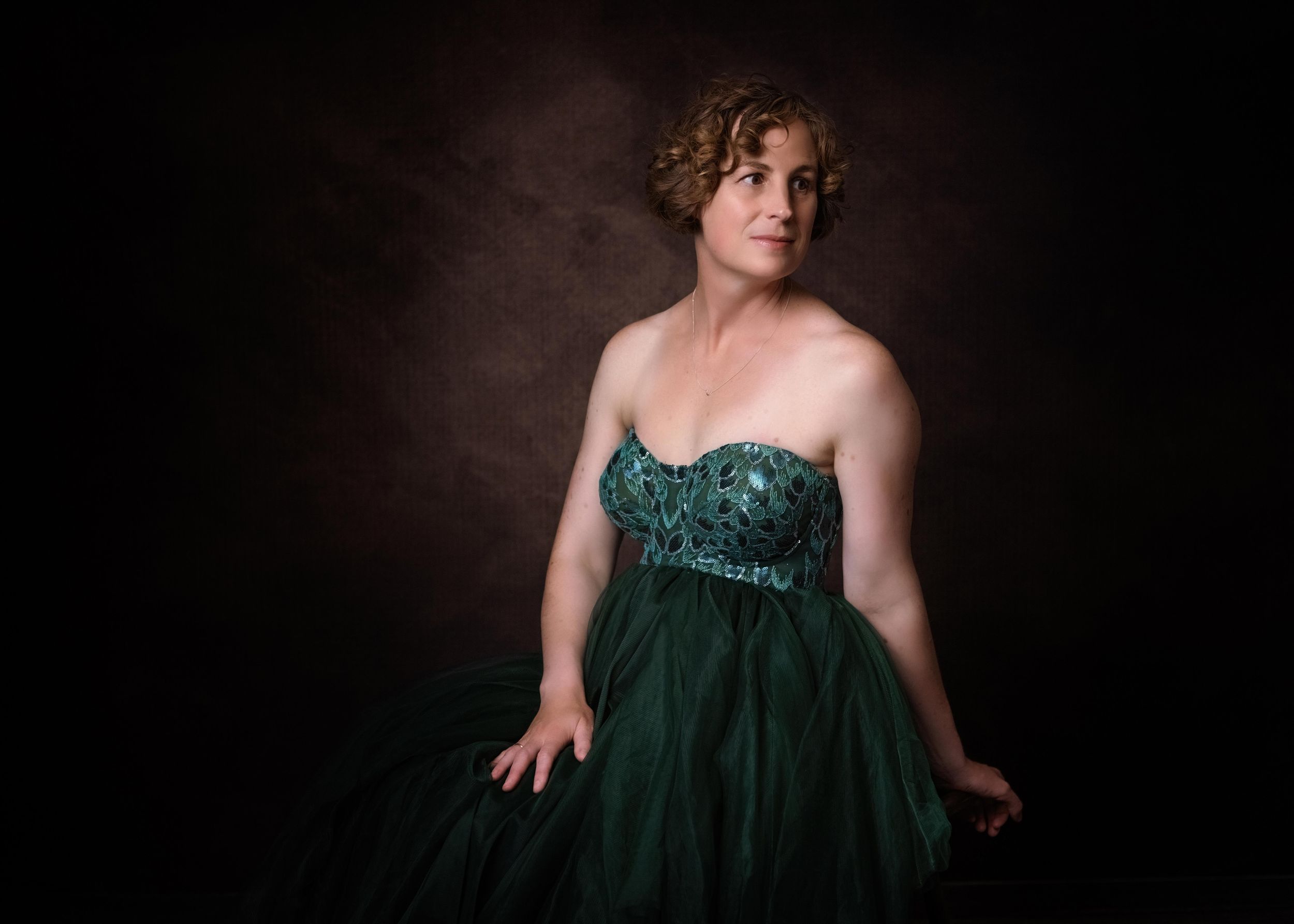 A person wearing an emerald green sequined formal gown poses against a dark backdrop in a studio portrait.