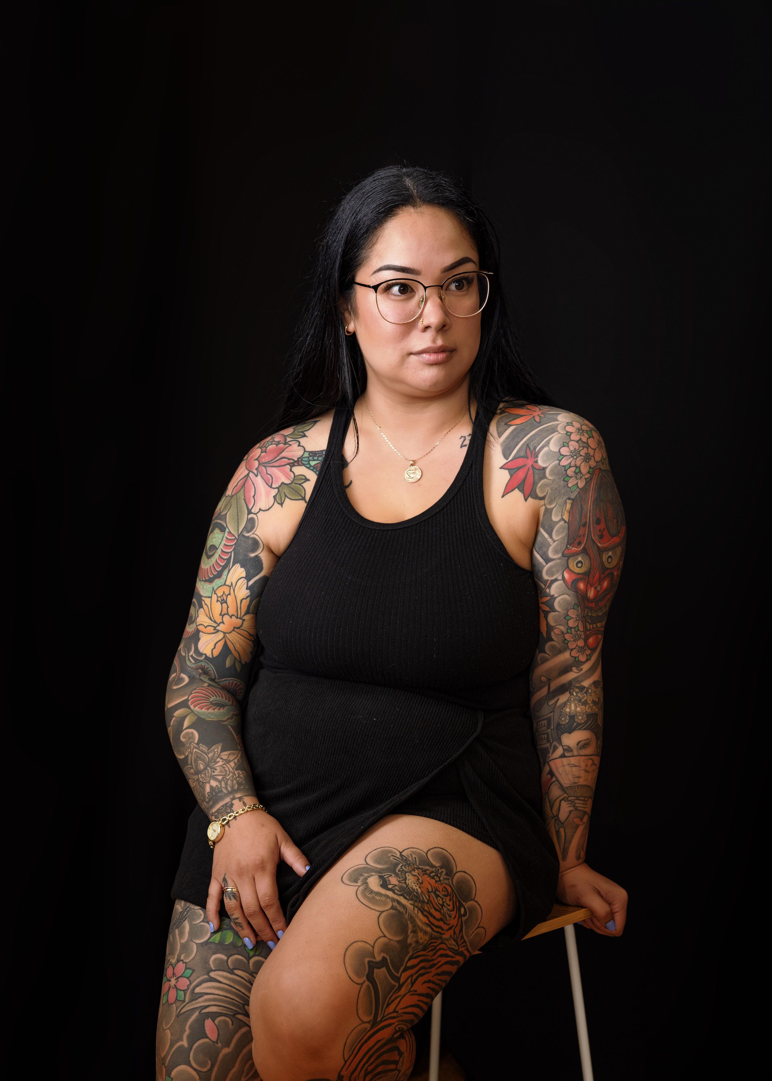 Studio portrait against black background showing person in black tank top with colorful sleeve tattoos striking dramatic pose.