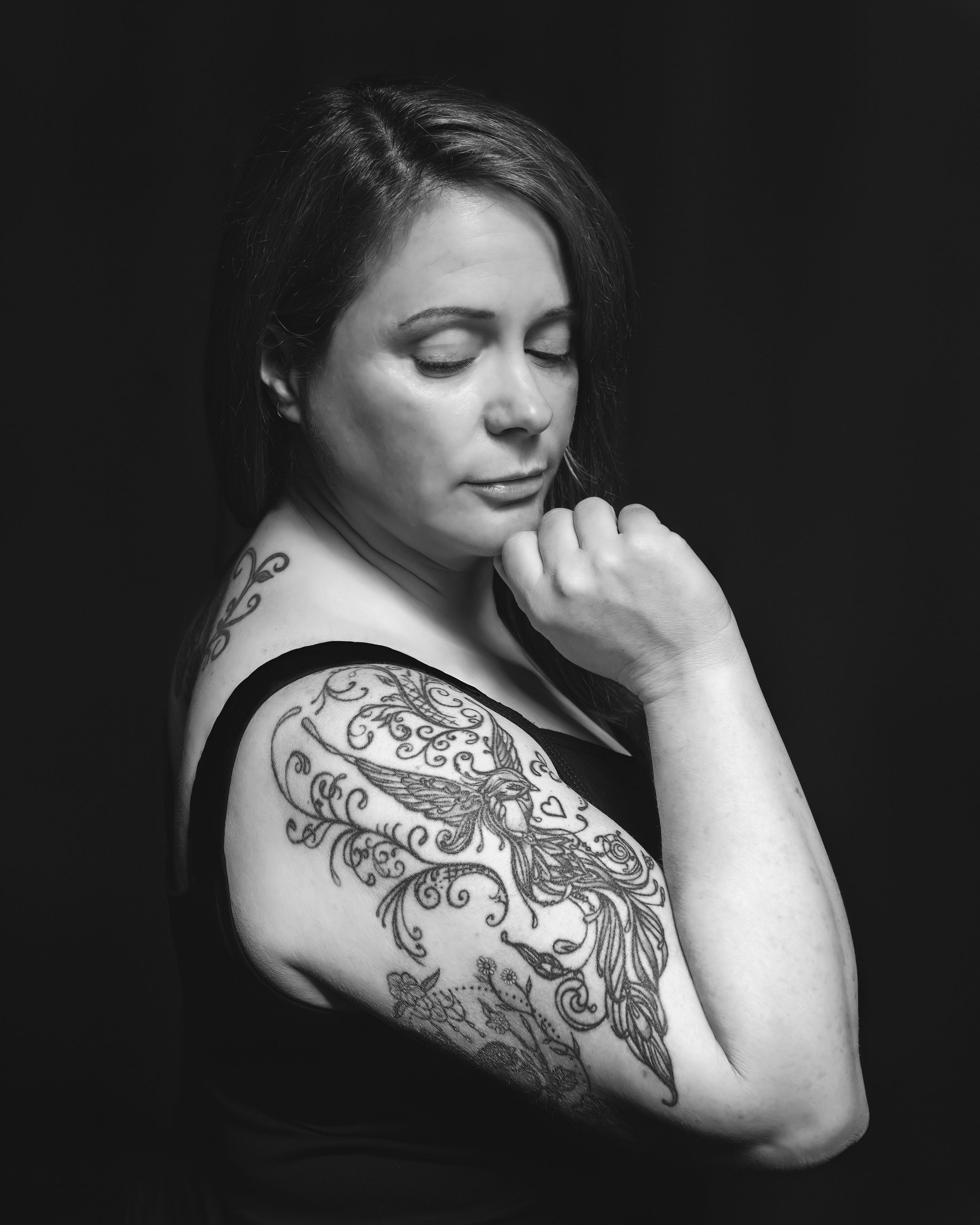 Black and white portrait showing a person with eyes closed wearing a floral and scrollwork shoulder tattoo.