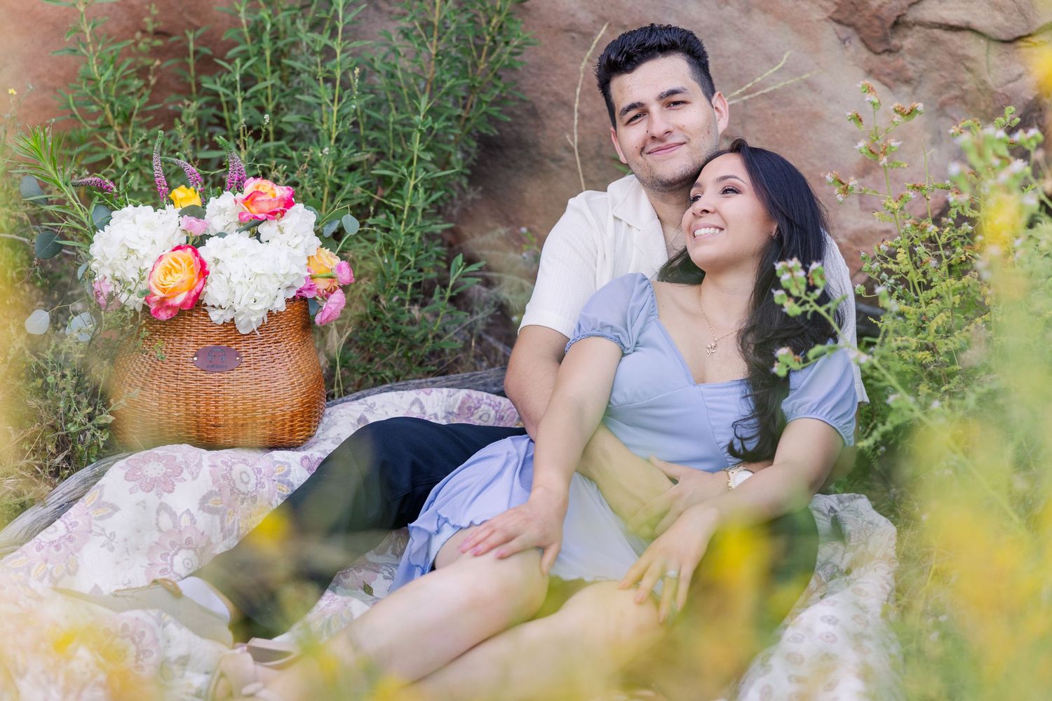 A romantic couple sits outdoors near a woven basket of colorful flowers among greenery and rocks. In Simi Valley CA