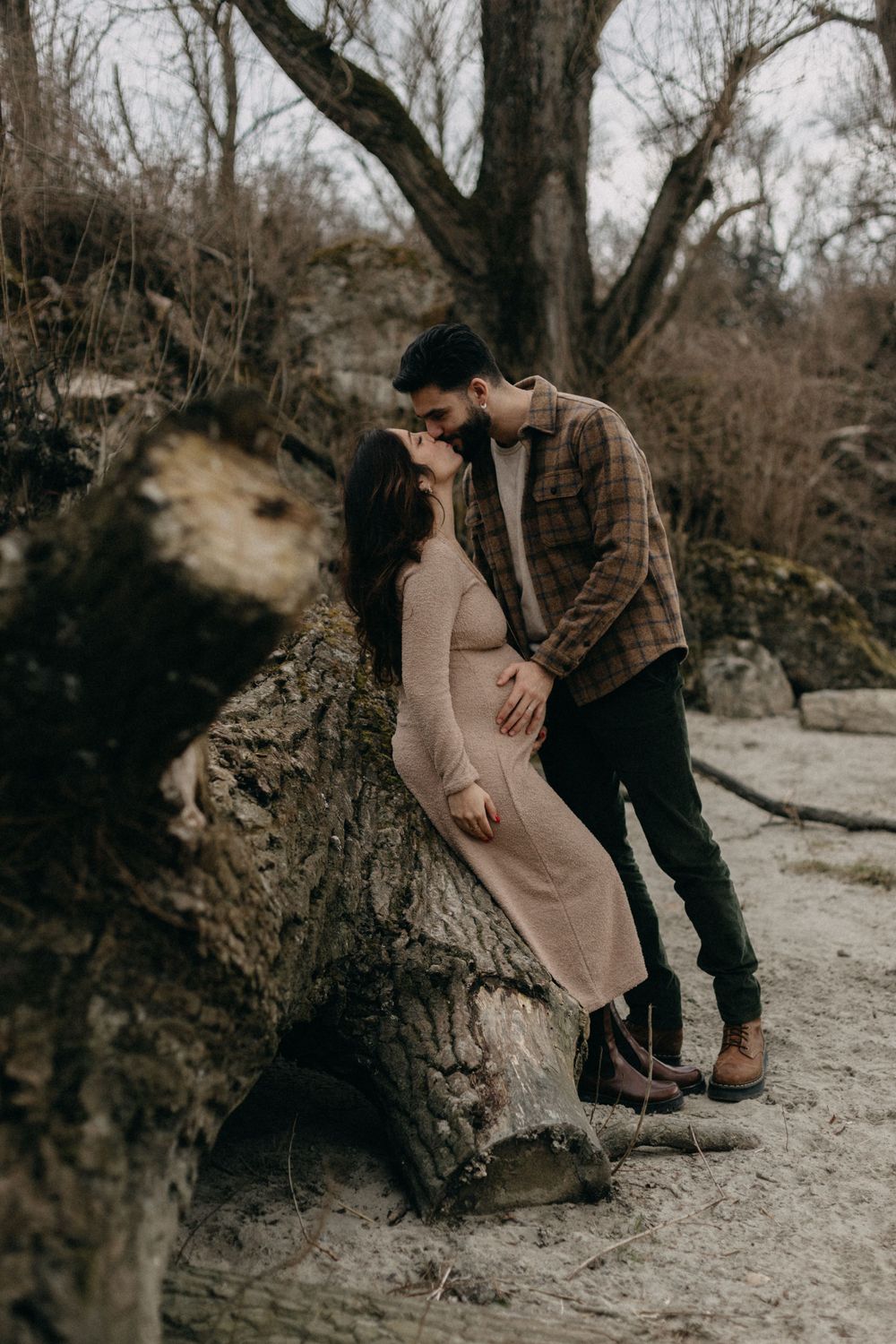 Romantic maternity portrait against fallen tree trunk in winter landscape.