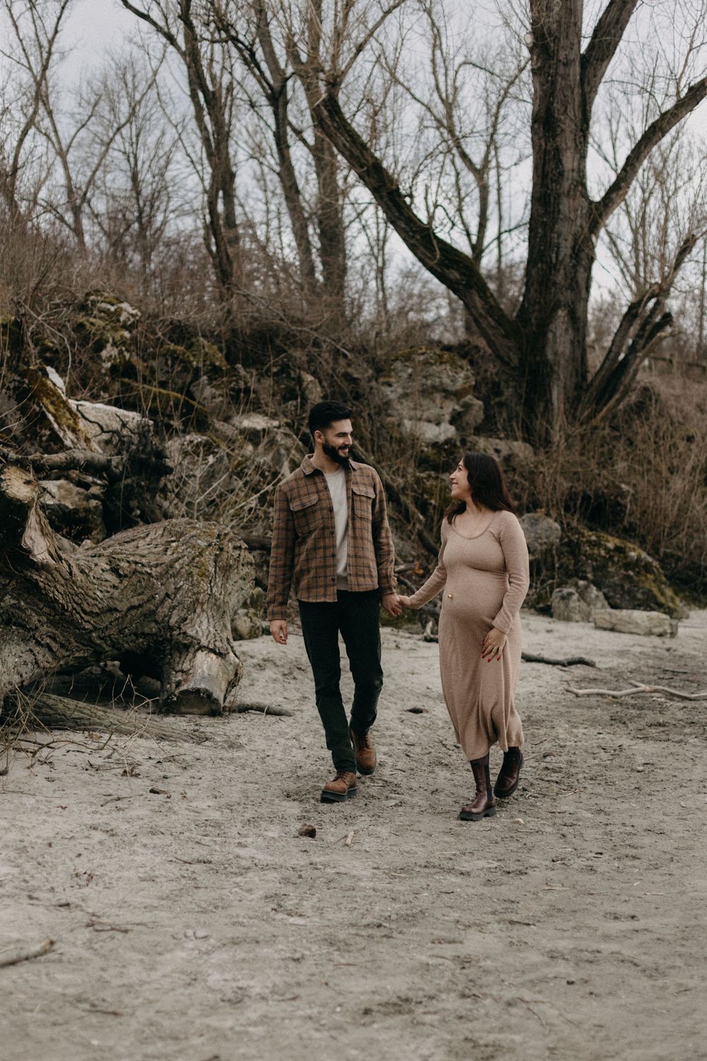 Couple takes romantic winter walk along sandy path with bare trees in background.