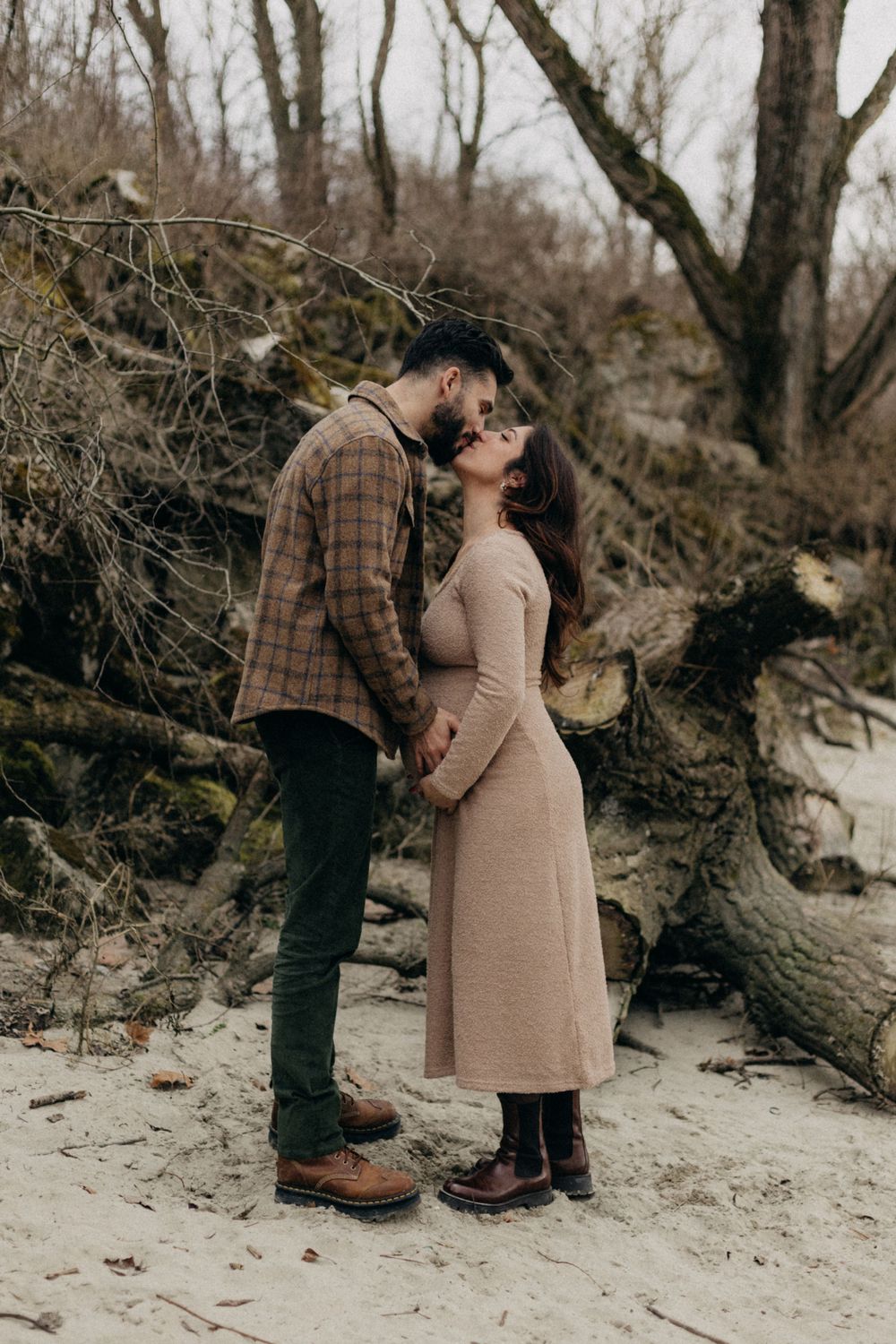 Intimate winter maternity portrait with couple standing on snowy ground near fallen trees.