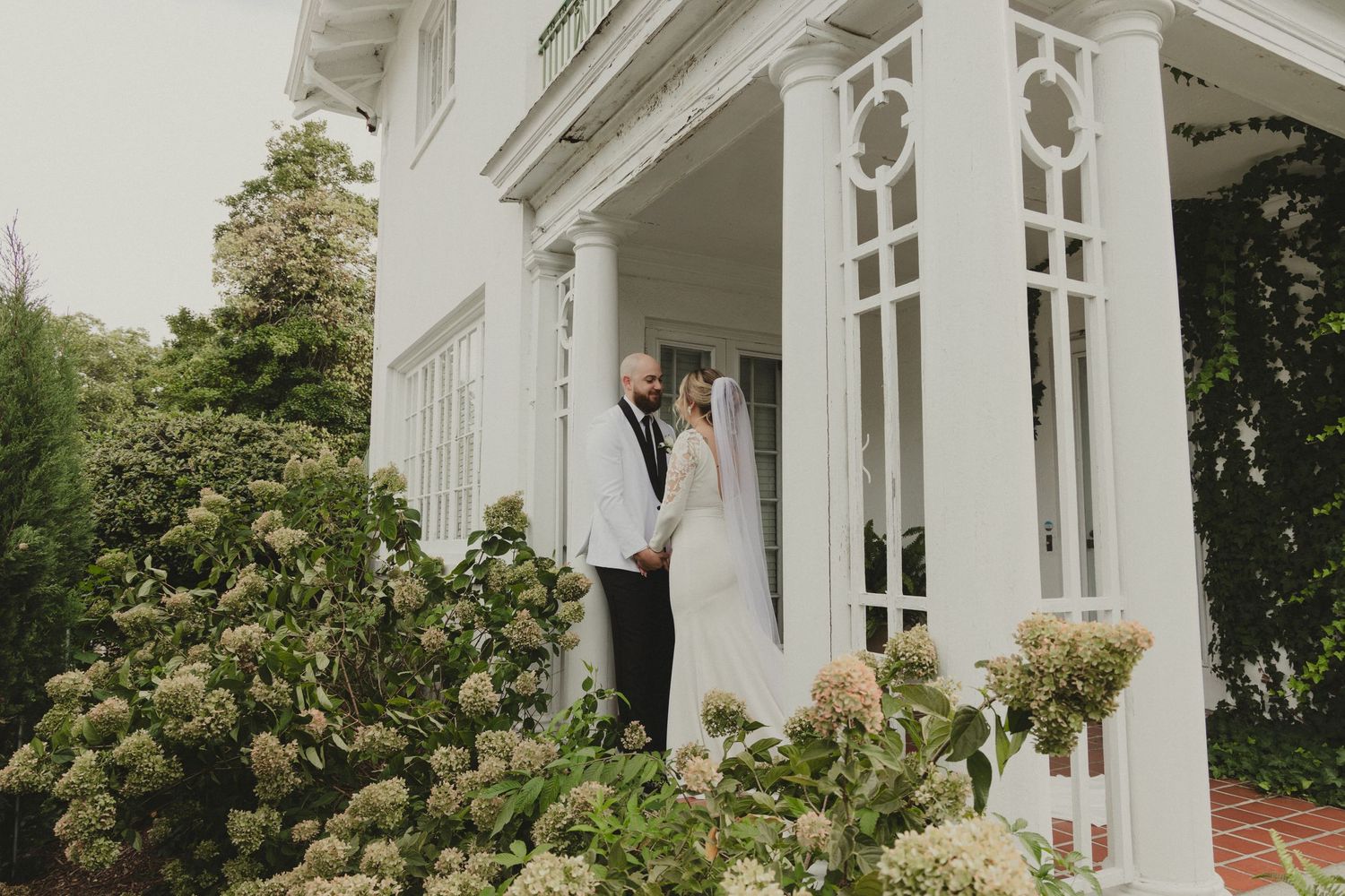 A couple stands in an elegant white porch with decorative columns surrounded by blooming hydrangea bushes.