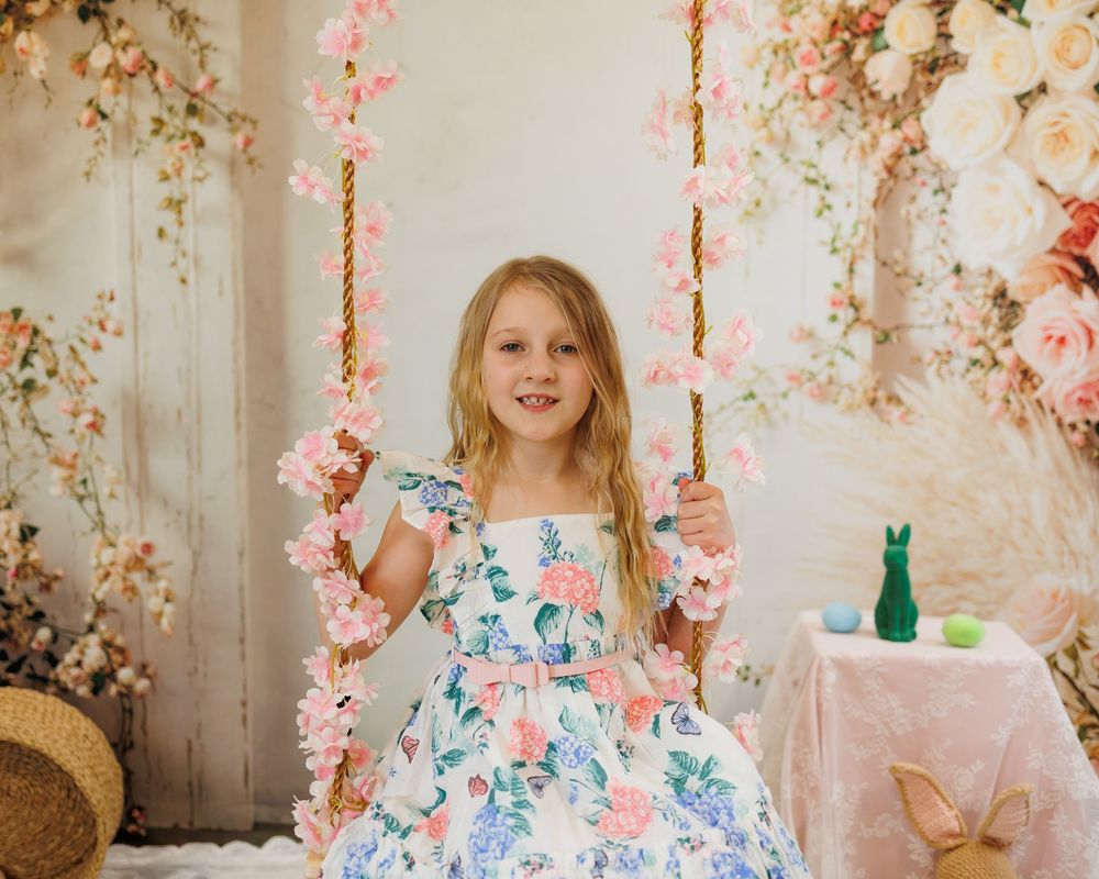Young girl in floral dress sitting on a flower-decorated swing in a spring-themed photo studio setting.