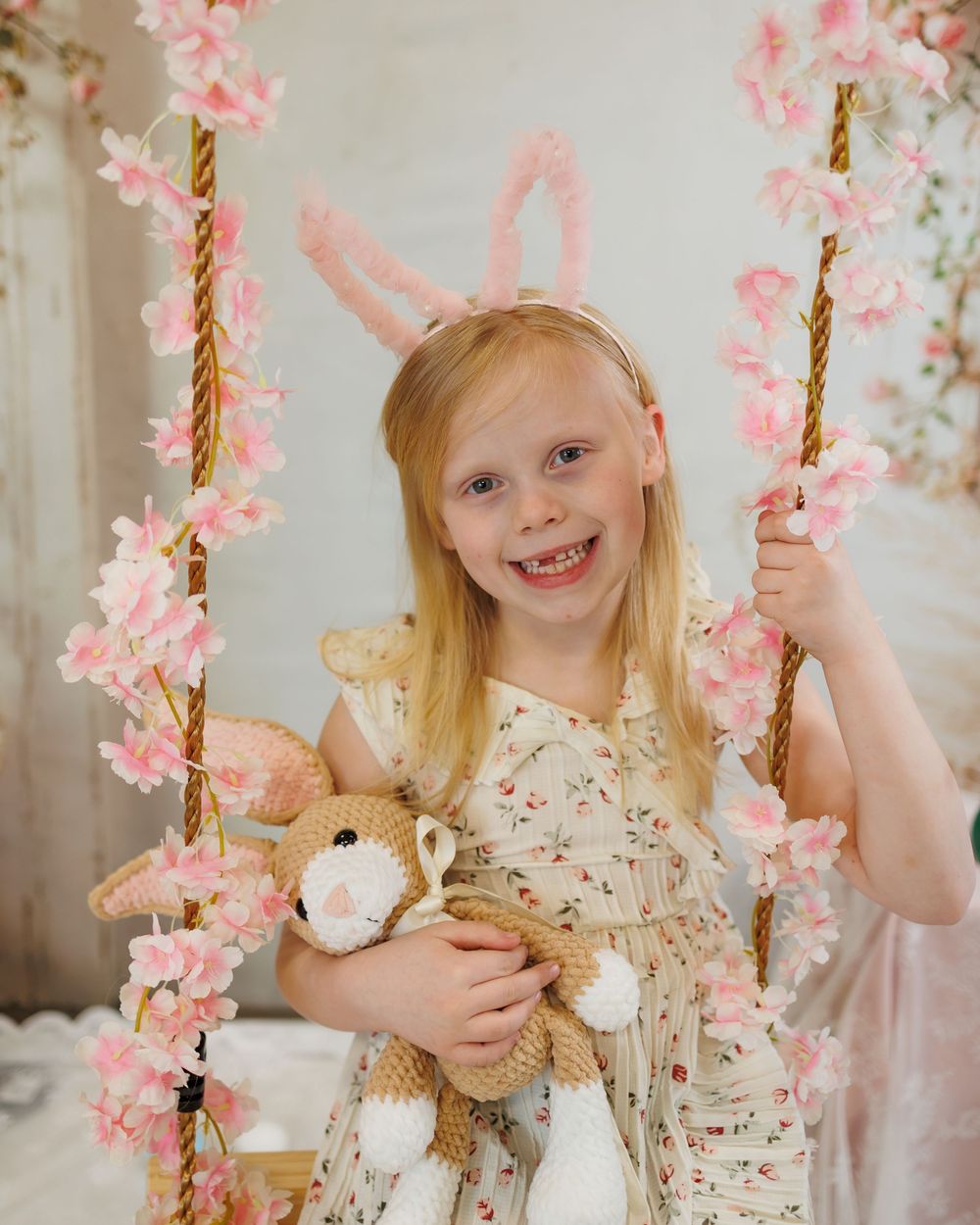 A smiling girl with bunny ears holds a stuffed rabbit on a floral swing with pink cherry blossoms.