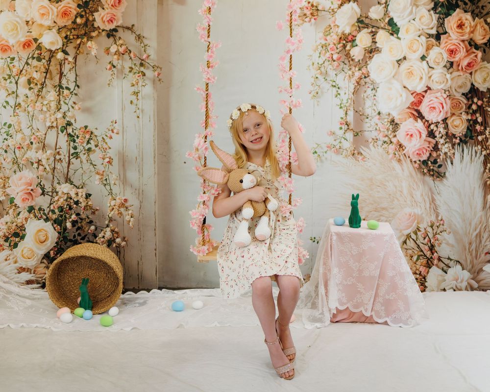 A young girl in a white dress and gold crown sits on a floral swing holding a teddy bear in a pastel spring setting.