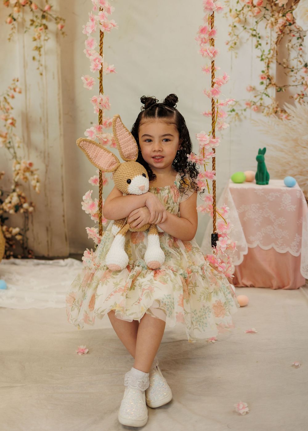 Young girl in floral dress sits on flower-decorated swing holding stuffed bunny toy in pastel Easter-themed photo setting.