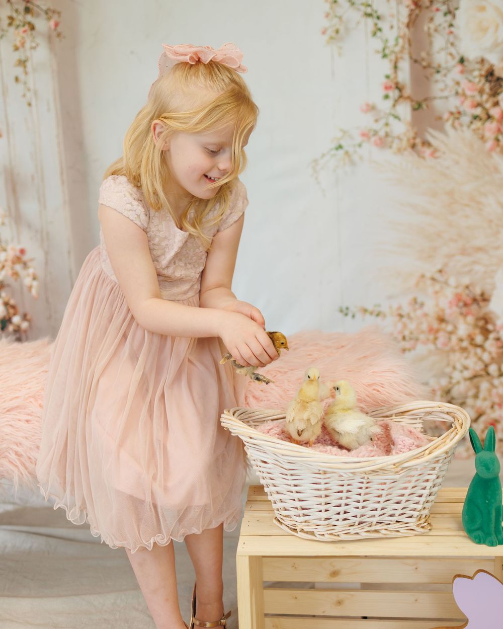 A child in a pink tulle dress bending down to look at a white basket containing a yellow duckling.