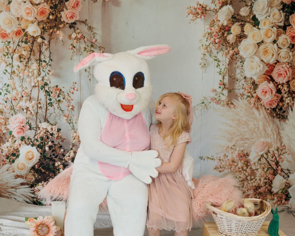 Whimsical Easter scene with large white bunny costume against floral backdrop of pink roses and spring decorations.