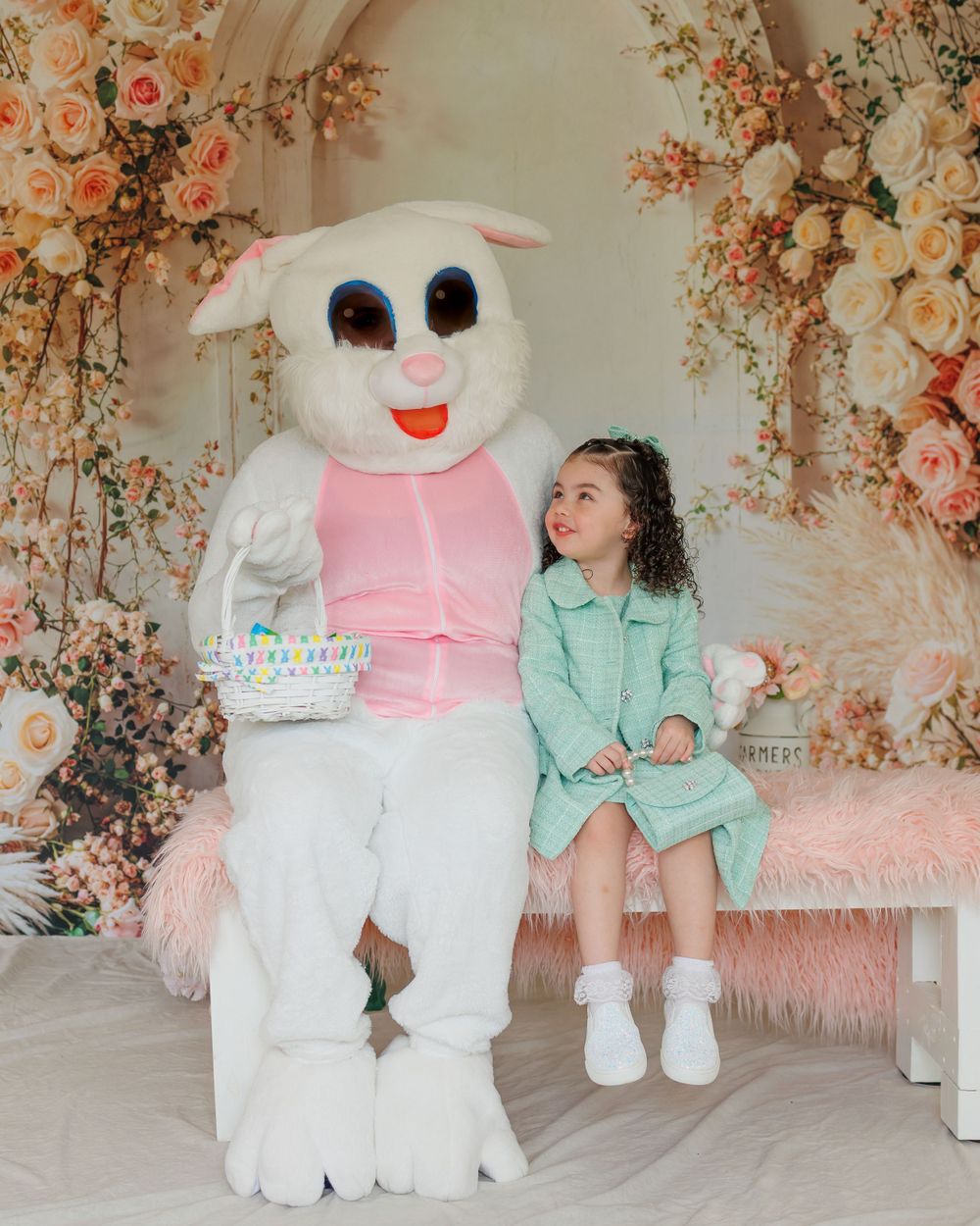 A large white Easter bunny mascot in pink vest sits next to a child wearing mint green on a floral backdrop.