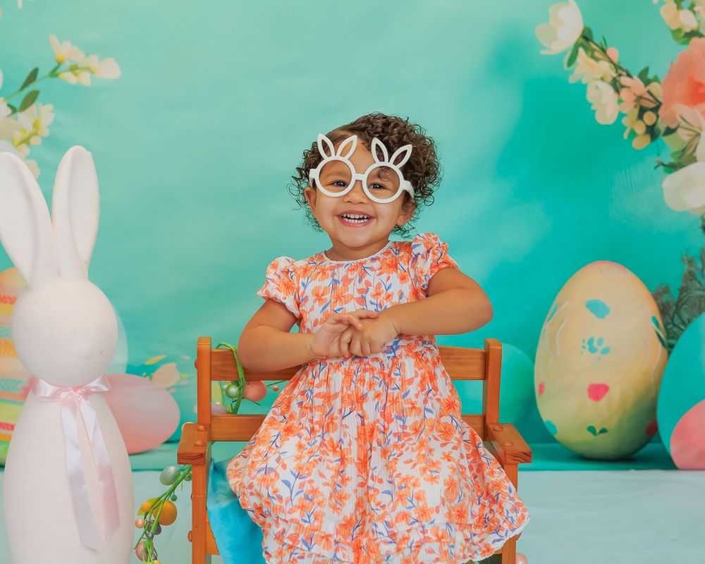 Easter photo setup with flowers, wooden chair, decorative bunny and pastel eggs against turquoise background.