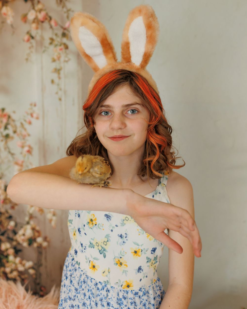 A person in a floral dress and bunny ears headband poses with a small hamster or gerbil in a springtime setting.