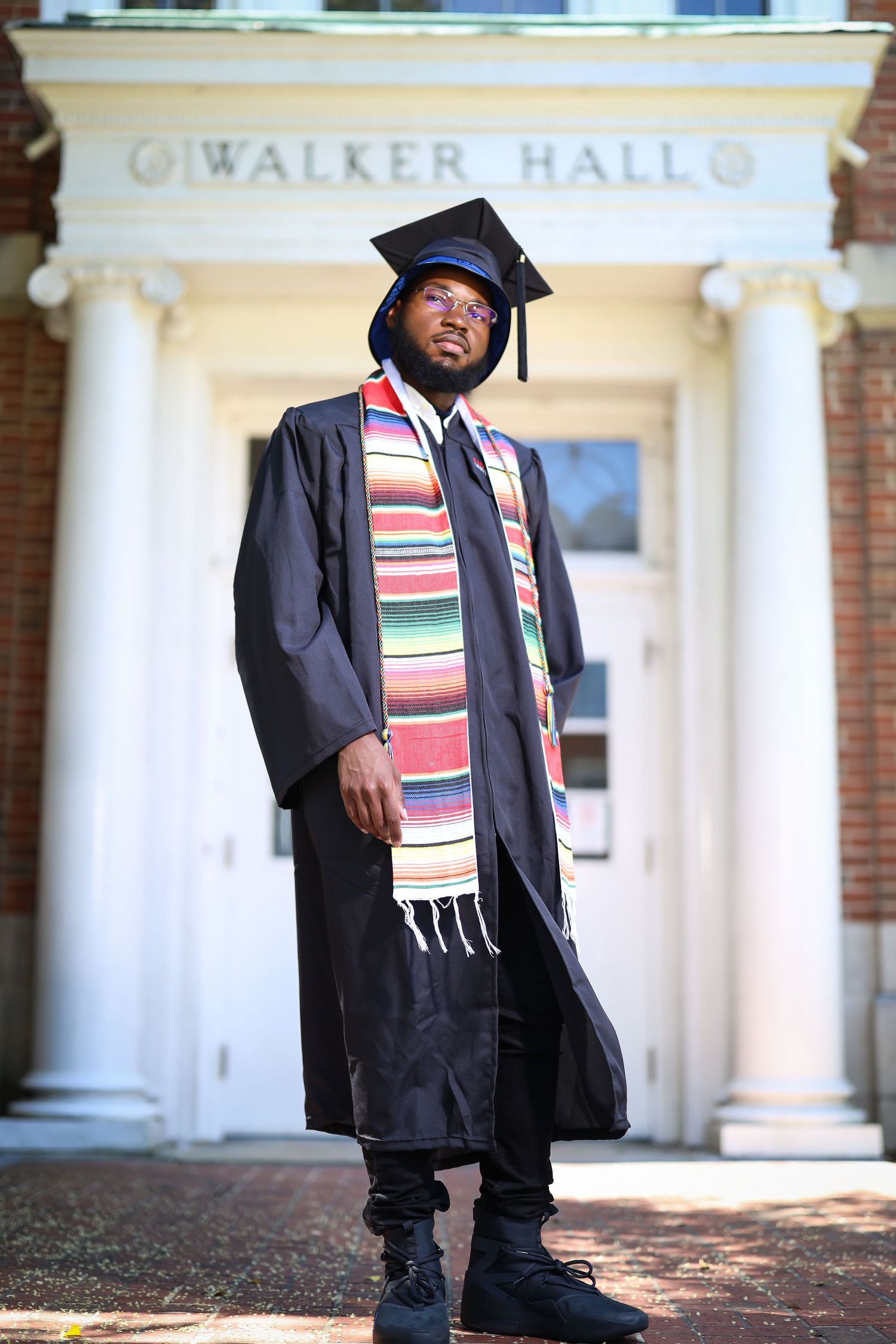 Graduate in cap and gown wearing colorful stole standing in front of Walker Hall with classical columns.