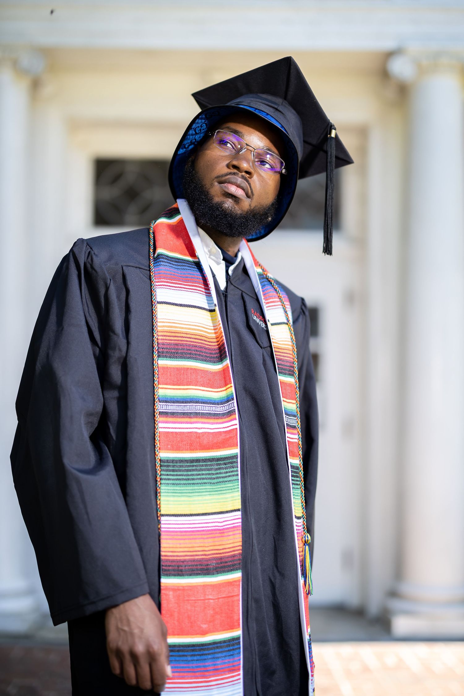 Graduate wearing traditional black cap and gown with vibrant striped stole against white pillared background.